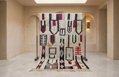 A large woven area rug with a geometric tribal pattern in cream, black, red, and navy, hanging from a wooden display stand in a minimalist beige hallway.