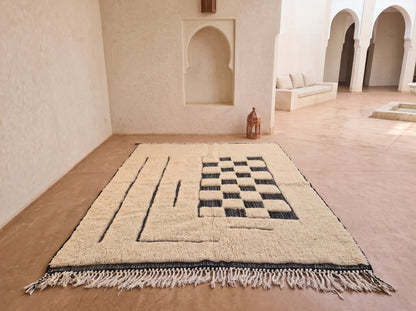 A rectangular wool rug with a cream and black geometric pattern and fringe, displayed on a terracotta floor in a Moroccan-style courtyard.
