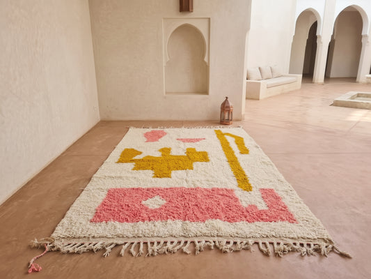 A rectangular woven rug with fringe, featuring a cream base with pink and mustard yellow geometric patterns, displayed on a terracotta floor in a Moroccan-style room with arched alcoves.