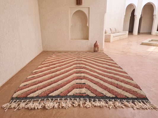 A large rectangular Moroccan-style rug with a chevron pattern in terracotta, beige, and dark charcoal, laid out on a terracotta floor in a minimalist, sunlit room with arched alcoves.