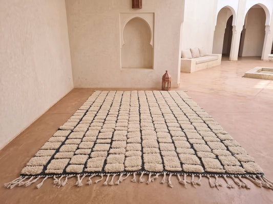 A large rectangular area rug with a textured, woven block pattern in cream and black, laid out on a terracotta tiled floor in a Moroccan-style room with arched alcoves.