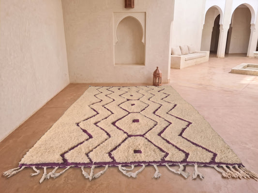 A rectangular area rug with a cream-colored shaggy texture and a purple geometric zigzag pattern, displayed on a terracotta tiled floor in a Moroccan-style room with arched alcoves.