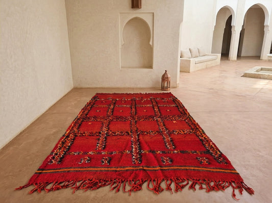 A red Moroccan-style woven rug with geometric patterns and fringe, displayed on a sandy floor in a minimalist, sunlit room with arched alcoves.