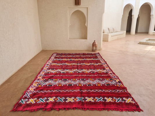 A rectangular Moroccan-style rug with a vibrant red base and intricate multicolor geometric patterns, displayed on a light beige stone floor in a traditional courtyard.