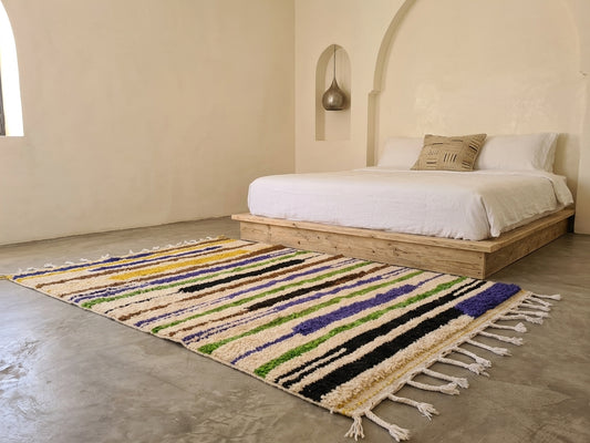 A colorful striped woven area rug with white fringe, styled on a concrete floor in front of a wooden platform bed with white bedding in a minimalist, warm-toned room.