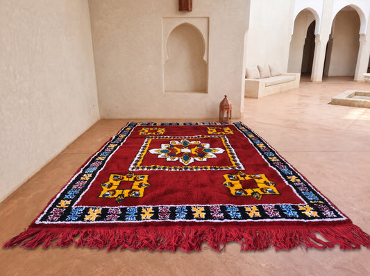 A large red Moroccan-style area rug with intricate multicolor geometric patterns and fringed edges, displayed on a terracotta floor in a sunlit Moroccan courtyard with white stucco walls and arches.