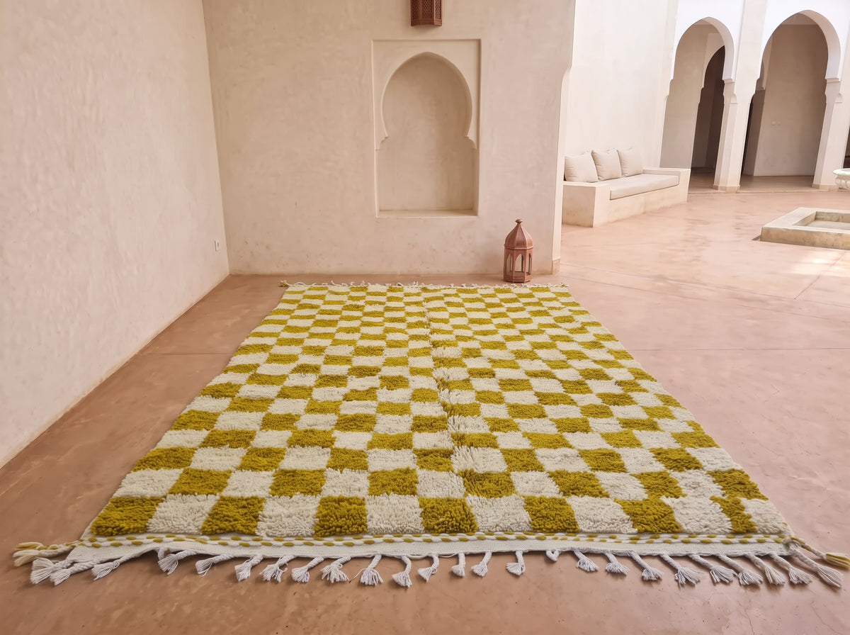 A large rectangular area rug with a yellow and off-white checkerboard pattern and fringe along one end, displayed on a terracotta floor in a Moroccan-style courtyard with arched alcoves in the background.