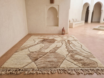 A large Moroccan-style area rug with a geometric tribal pattern in beige, brown, and cream tones, laid out on a terracotta floor in a minimalist, sunlit room with arched alcoves.