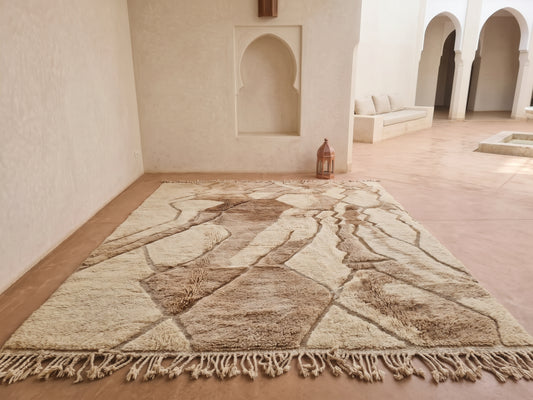 A large Moroccan-style area rug with a geometric tribal pattern in beige, brown, and cream tones, laid out on a terracotta floor in a minimalist, sunlit room with arched alcoves.