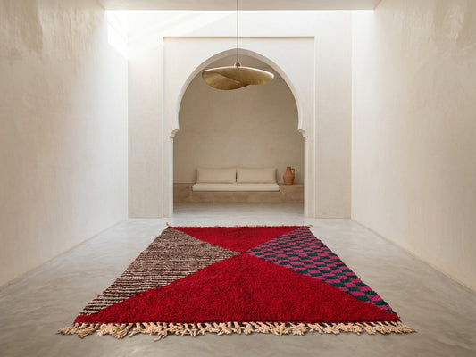 A Moroccan-style area rug with a bold geometric pattern in red, beige, and blue, displayed on a concrete floor in a minimalist, cream-colored room with an arched doorway in the background.