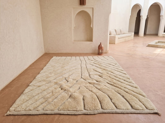 A large rectangular beige rug with a raised geometric leaf-like pattern, laid out on a terracotta-colored floor in a minimalist Moroccan-style courtyard with arched alcoves and a small lantern.