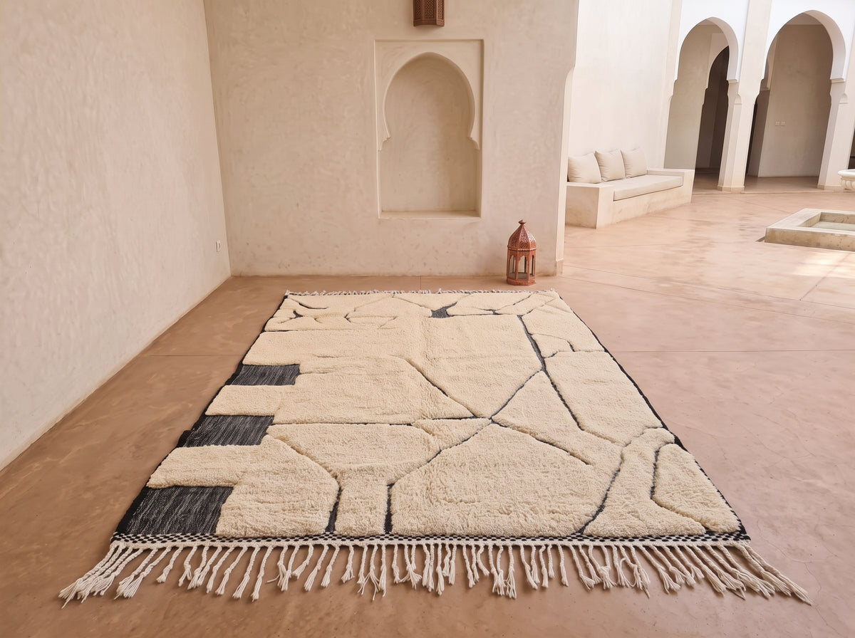A large rectangular area rug with a cream and black geometric pattern and fringe, displayed on a terracotta floor in a Moroccan-style courtyard with arched alcoves.