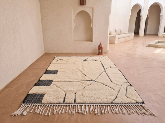 A large rectangular area rug with a cream and black geometric pattern and fringe, displayed on a terracotta floor in a Moroccan-style courtyard with arched alcoves.