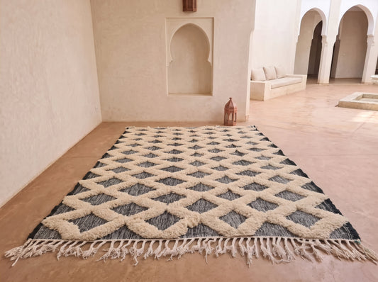 A large rectangular Moroccan-style rug with a cream and dark gray geometric diamond pattern and fringe, displayed on a terracotta floor in a minimalist, sunlit room with arched alcoves.