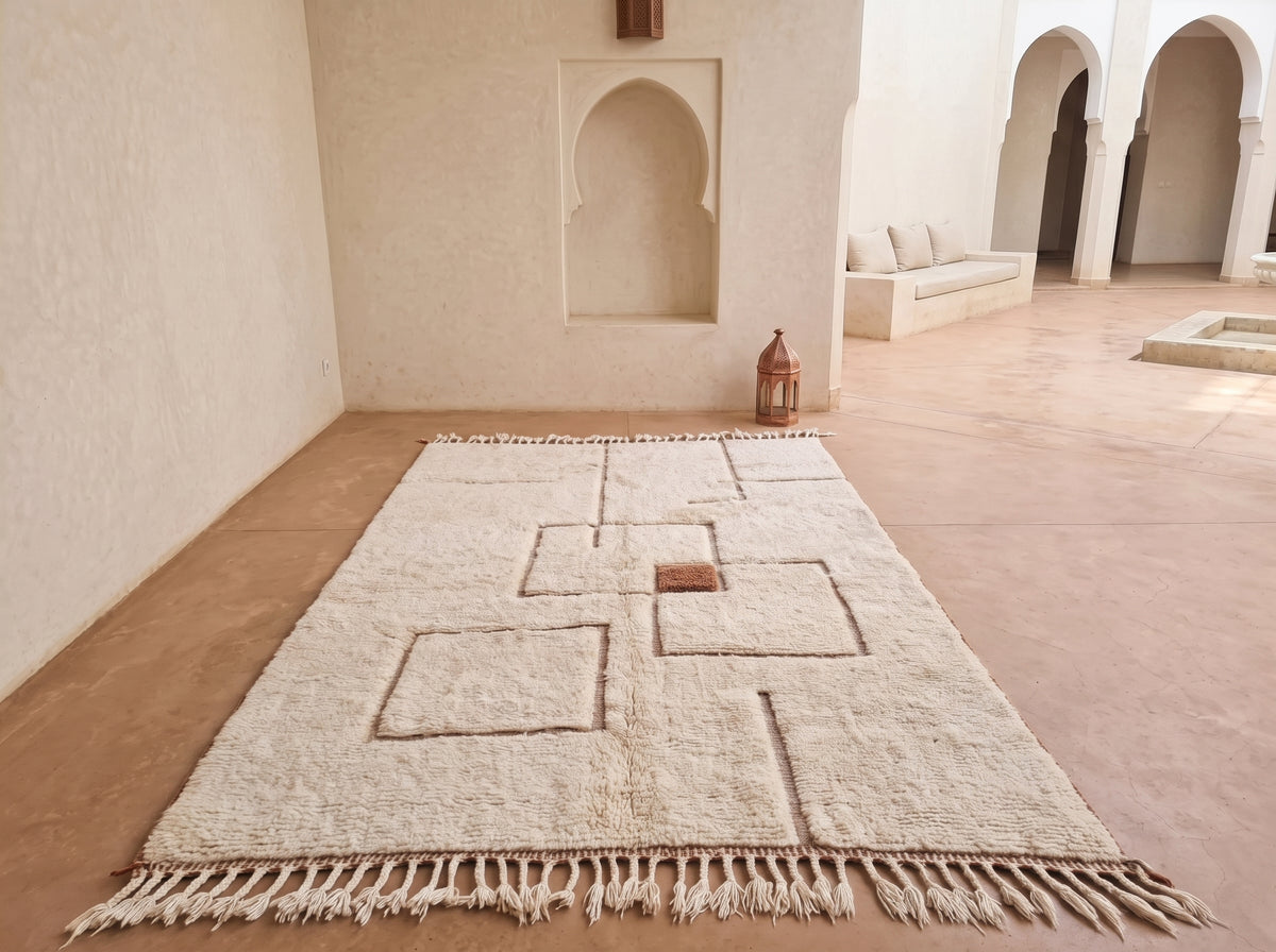 A large off-white wool rug with geometric raised patterns and fringe, displayed on a terracotta-colored floor in a Moroccan-style courtyard with arched alcoves.