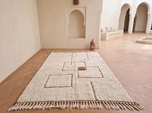 A large off-white wool rug with geometric raised patterns and fringe, displayed on a terracotta-colored floor in a Moroccan-style courtyard with arched alcoves.