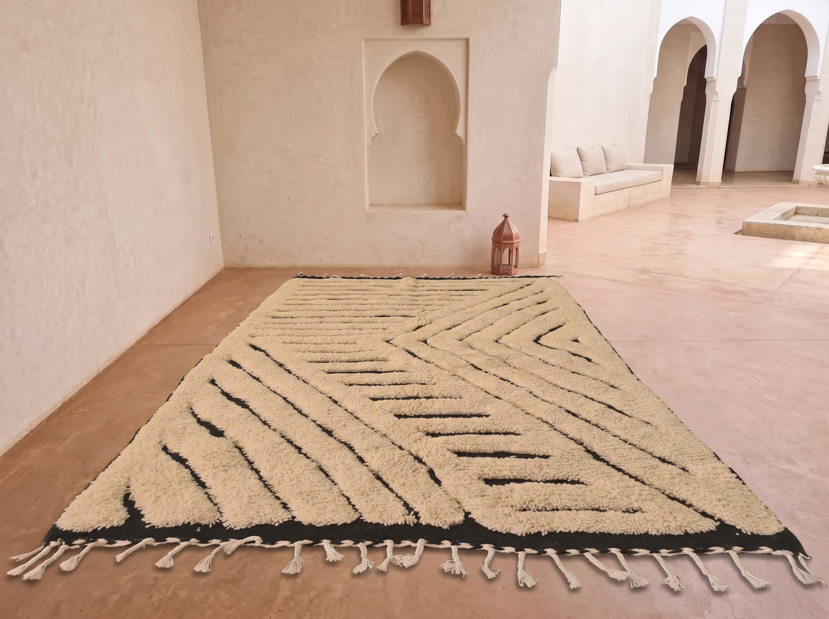 A large rectangular area rug with a cream and black geometric pattern and tasseled fringe, displayed on a terracotta tiled floor in a Moroccan-style room with arched alcoves.