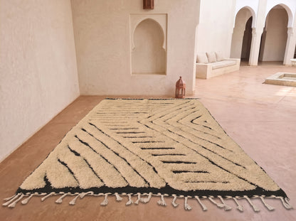 A large rectangular area rug with a cream and black geometric pattern and tasseled fringe, displayed on a terracotta tiled floor in a Moroccan-style room with arched alcoves.