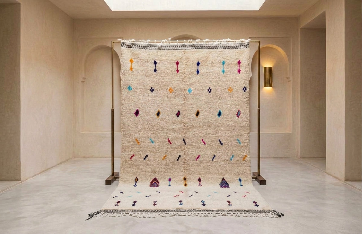 A large cream-colored Moroccan-style rug displayed on a black metal stand in a minimalist beige gallery space. The rug features colorful embroidered geometric patterns and tassels, with a matching rug laid out on the floor in front.