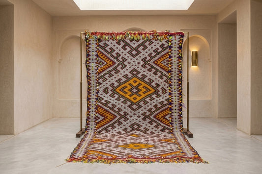 A large woven Moroccan-style rug displayed on a black metal stand in a minimalist beige hallway. The rug features a complex geometric pattern with a central diamond motif in yellow, orange, and brown on a cream background, surrounded by red, blue, and green accents.
