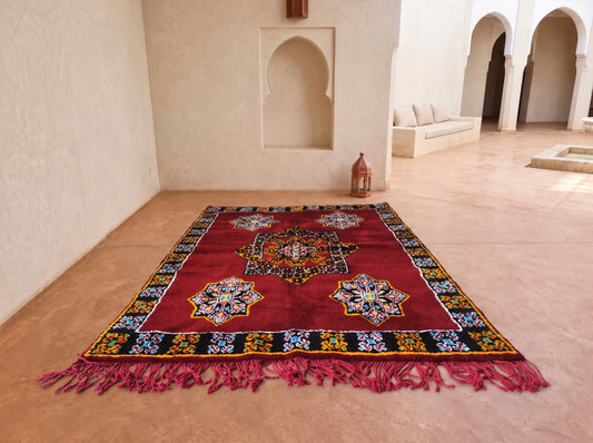 A rectangular Moroccan-style area rug with a deep red field, intricate multicolor medallion motifs, and a patterned border, displayed on a terracotta tiled floor in a sunlit courtyard with cream walls and arches.