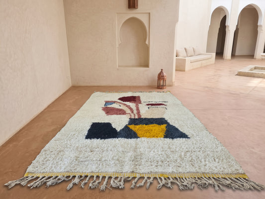 A large white Moroccan wool rug with a colorful geometric pattern and fringed edges, displayed on a terracotta tiled floor in a minimalist, sunlit room with arched alcoves.