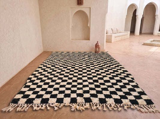 A large black and off-white checkered rug with tasseled fringe, laid out on a terracotta-colored floor in a Moroccan-style room with arched alcoves.
