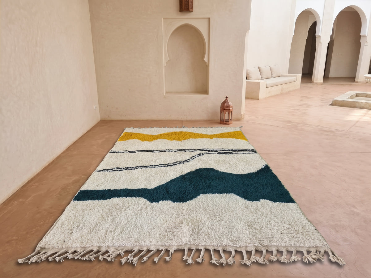 A rectangular shag area rug with a cream base and wavy horizontal stripes in mustard yellow, dark teal, and navy, displayed on a terracotta tiled floor in a Moroccan-style room with arched alcoves.