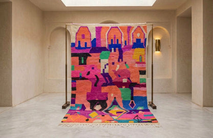 A large colorful woven rug displayed on a metal stand in a minimalist beige gallery space with arched walls and skylights.