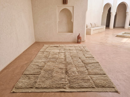 A large rectangular beige area rug with a textured, patchwork-like woven pattern, displayed on a terracotta-colored floor in a Moroccan-style courtyard with white stucco walls and arches.