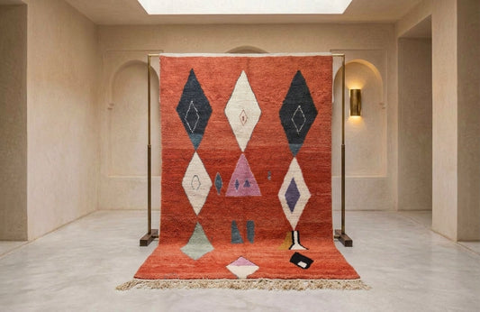 A large Moroccan-style woven rug displayed on a metal stand in a minimalist beige gallery space. The rug features a terracotta red-orange base with large geometric diamond and triangle motifs in cream, navy blue, light purple, and pale green.