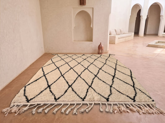 A large rectangular area rug with a black diamond lattice pattern on an off-white background, featuring fringe along the bottom edge, displayed on a terracotta floor in a Moroccan-style room with arched alcoves.