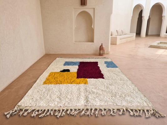 A large white shag rug with colorful geometric blocks and a fringed edge, displayed on a terracotta floor in a minimalist Moroccan-style room with arches and a bench.