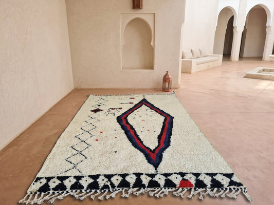 A Moroccan-style wool area rug with a cream base, featuring a large central diamond motif in red, blue, and black, and a black-and-white geometric border with white fringe tassels, displayed on a terracotta tiled floor in a Moroccan-style courtyard.