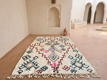 A rectangular Moroccan-style area rug with a cream base and colorful geometric patterns, displayed on a terracotta-colored floor in a Moroccan-style courtyard with arches and a bench in the background.
