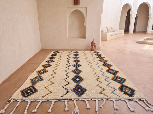 A large Moroccan-style area rug with a cream base and colorful geometric patterns, laid out on a terracotta tiled floor in a minimalist courtyard with white stucco walls and arches.