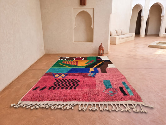 A colorful Moroccan-style area rug with cream fringe, displayed on a terracotta tiled floor in a minimalist, sunlit courtyard with arched walls.