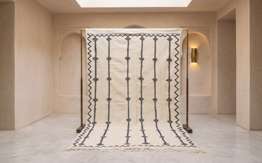 A large cream-colored wool rug with a black geometric pattern, displayed on a wooden stand inside a minimalist beige room with arched walls and warm lighting.
