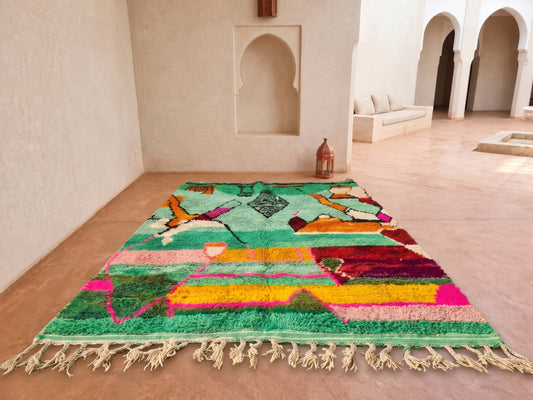 A vibrant Moroccan-style wool rug with multicolored geometric patterns and cream fringe, displayed on a terracotta floor in a minimalist, sunlit courtyard with arched walls.