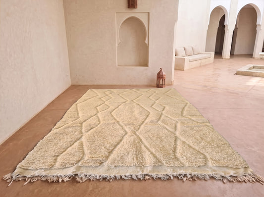 A large beige Moroccan-style rug with a raised geometric diamond pattern and fringed edges, displayed on a terracotta-colored floor in a minimalist, sunlit room with arched alcoves.