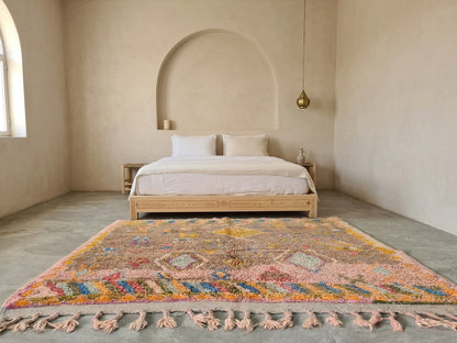 A large, colorful Moroccan-style shag rug with pink, orange, blue, and yellow patterns and matching tassels, styled on a concrete floor in a minimalist, warm-toned bedroom with a wooden bed.
