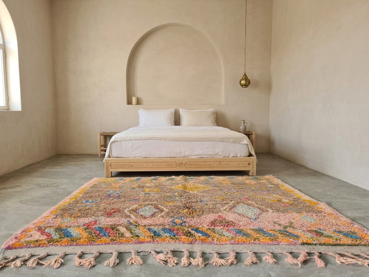 A large, colorful Moroccan-style shag rug with pink, orange, blue, and yellow patterns and matching tassels, styled on a concrete floor in a minimalist, warm-toned bedroom with a wooden bed.