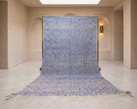A large blue and white patterned rug or textile panel hanging vertically in a minimalist beige interior space with arched wall openings and a light floor.