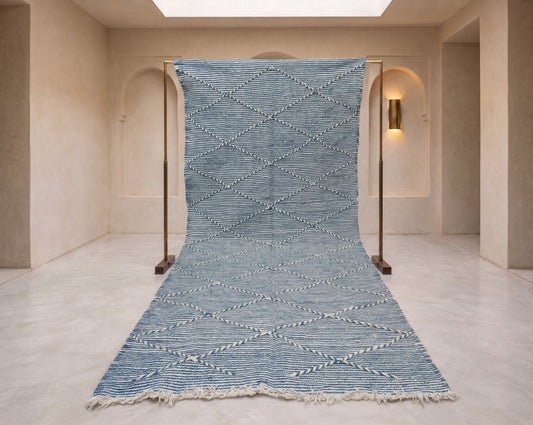 A long blue and white patterned rug displayed on a black metal stand in a minimalist beige room with arched walls and a skylight.