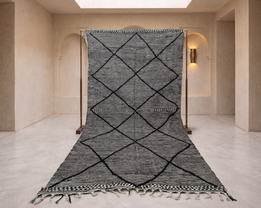 A large woven rug displayed on a wooden support frame in a minimalist beige interior space. The rug is charcoal gray with a black diamond lattice pattern and fringed edges.