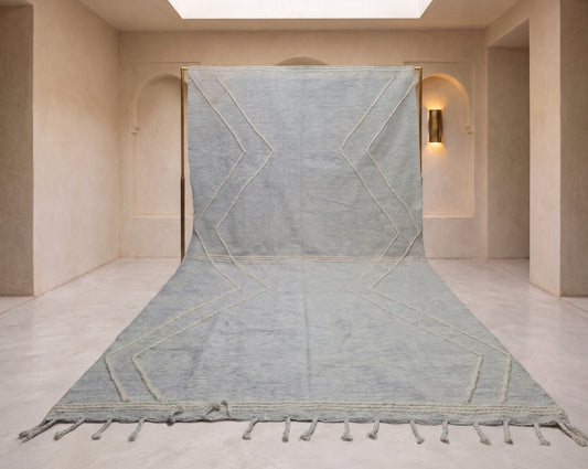 A large woven area rug in a muted sage green color with a geometric diamond pattern and tasseled fringe, displayed hanging from a wall in a minimalist beige room.