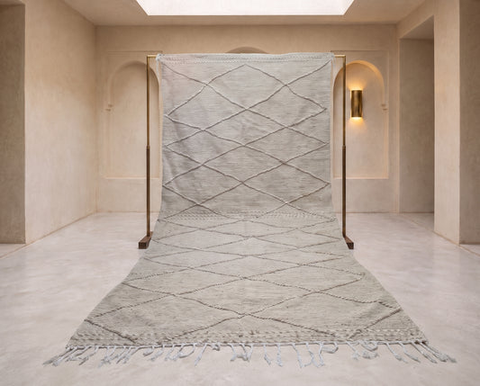 A large light gray woven rug with a diamond pattern and fringe, displayed on a wooden stand in a minimalist beige room.