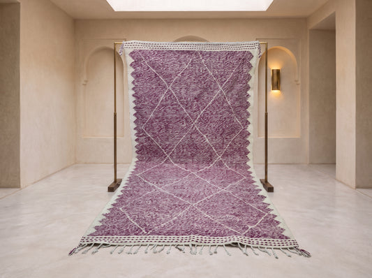 A purple and white patterned Moroccan-style rug displayed on a metal stand in a minimalist beige room with arched walls and a skylight.