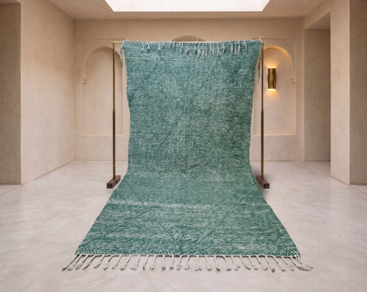 A large teal green woven rug with white fringe, displayed hanging from a wooden stand in a minimalist beige stone room.