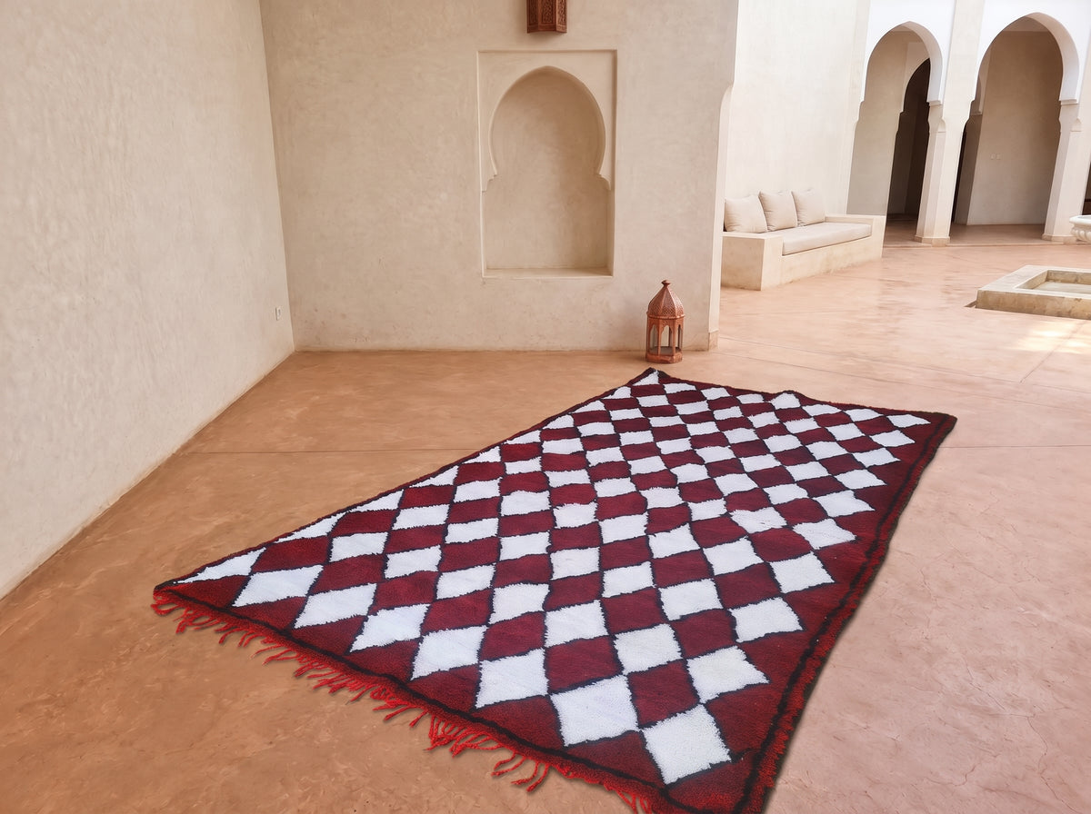 A rectangular area rug with a red and white diamond checkered pattern and red fringe, displayed on a terracotta tiled floor in a Moroccan-style courtyard with beige walls and arches.